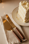Brushed gold cake server and knife with hardwood handles on cream linen napkin with white wedding cake in background