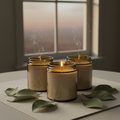 Three amber glass candle jars on cream linen runner with dried magnolia leaves and soft candlelight glow