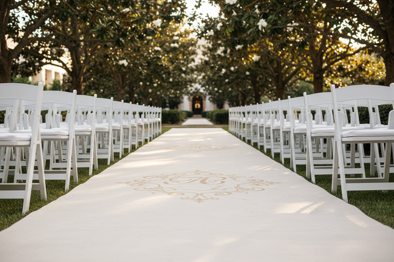 Ivory canvas aisle runner unrolling down garden courtyard aisle with white chairs and magnolia trees in golden afternoon light