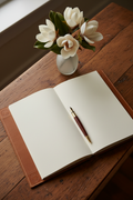 Saddle tan leather guest book open on vintage wood console with brass fountain pen and magnolia vase in warm amber light