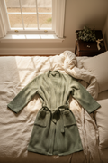 Sage green waffle-cotton robe laid flat on ivory linen bed with pearl earrings and eucalyptus bouquet